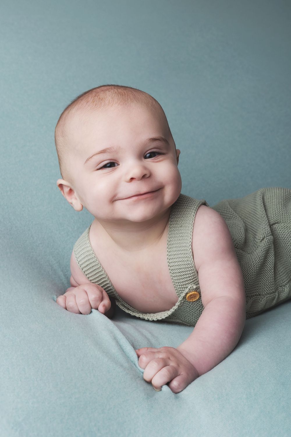 Happy baby in a sage green knit romper lies on tummy on soft blue backdrop during professional photo session.