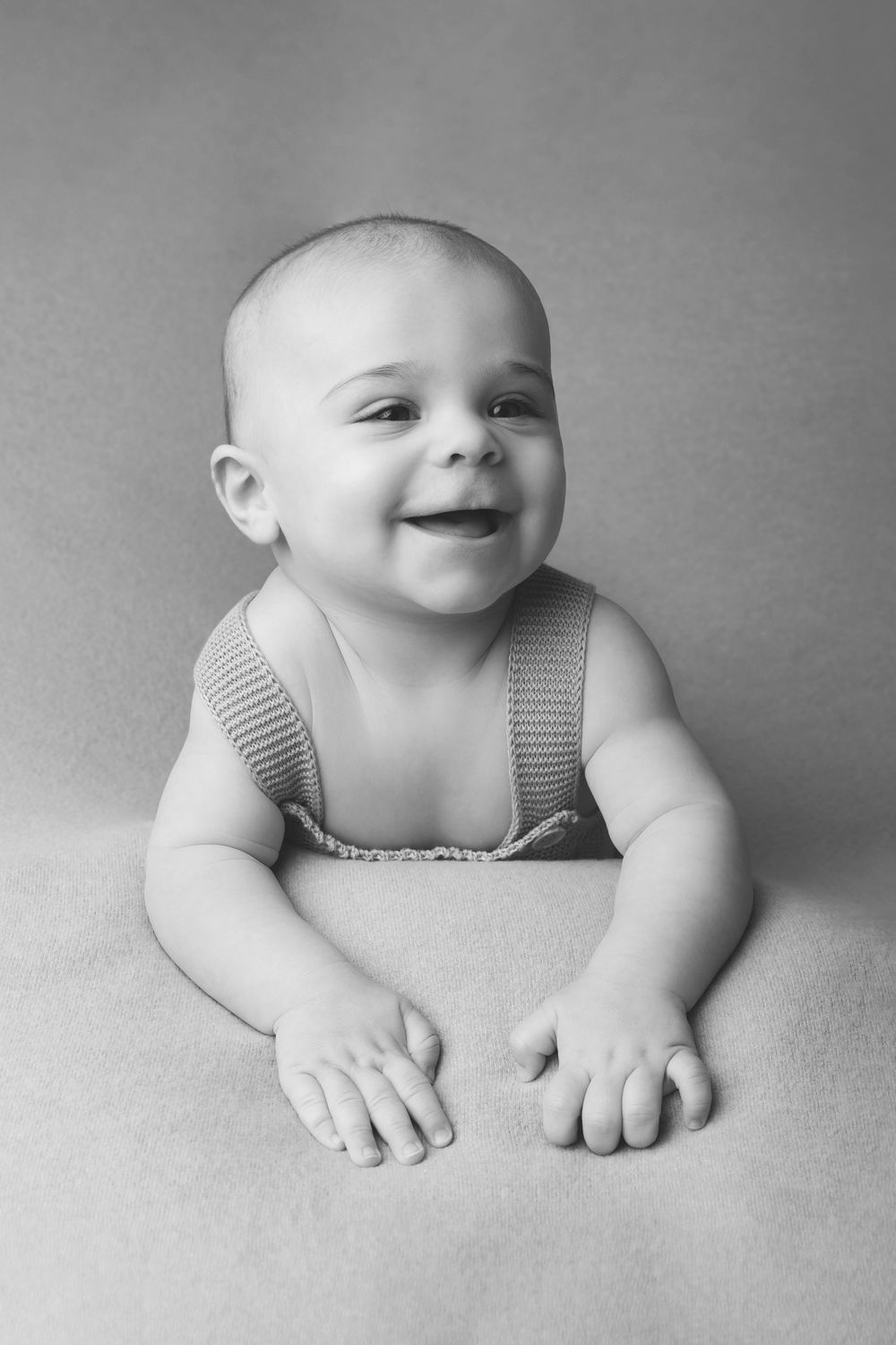 A black and white portrait of a smiling baby lying on their stomach wearing striped overalls.