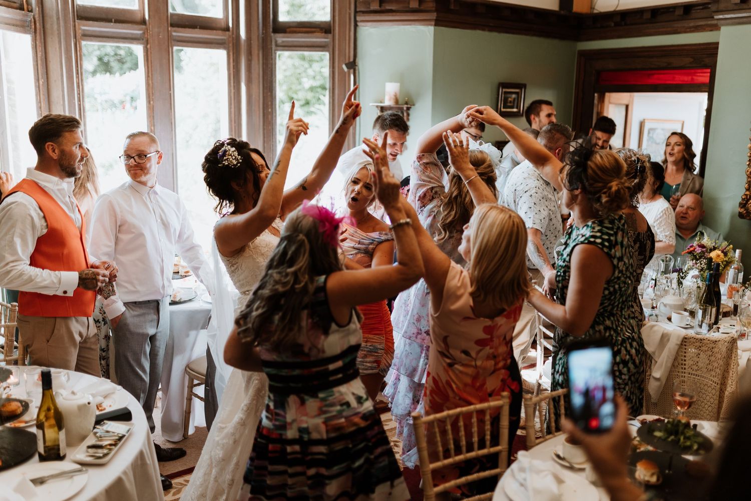 A group of wedding guests dancing and celebrating together in an indoor reception venue with wood trim windows.