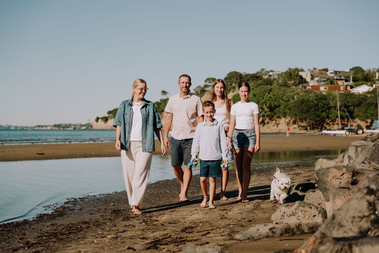 Group walking together with a dog along a scenic coastal beach path as the sun sets behind hills in the background.