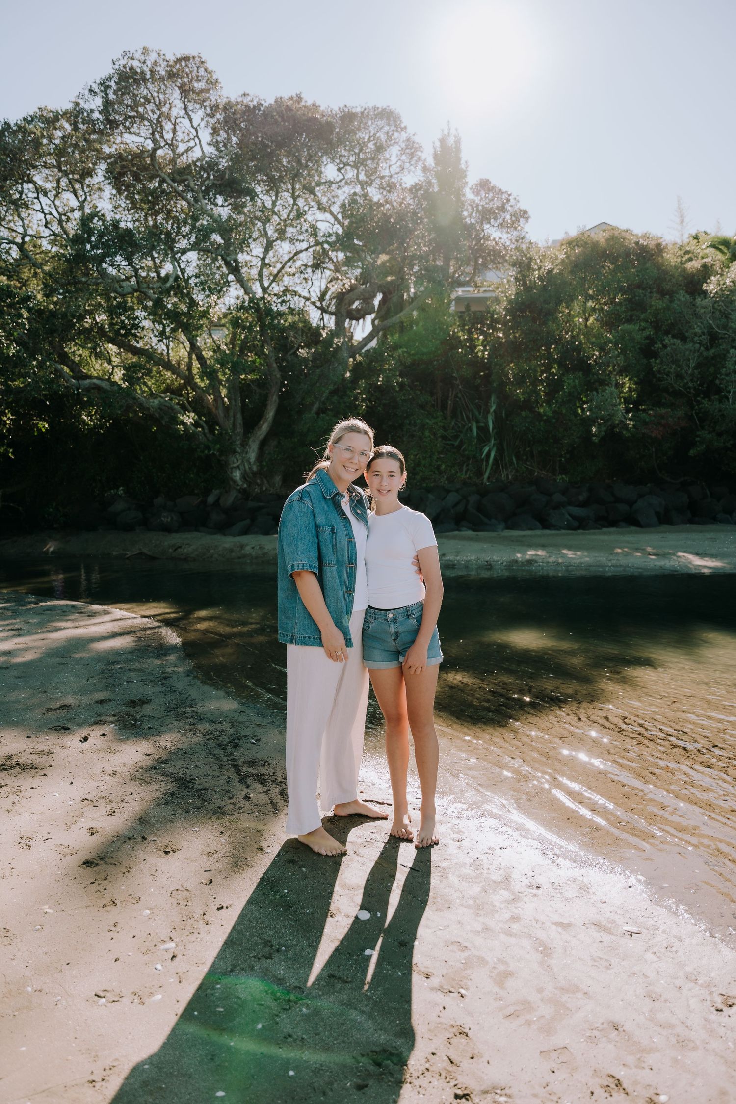 A couple stands together on a wooden dock by a tranquil river surrounded by lush trees on a sunny day.