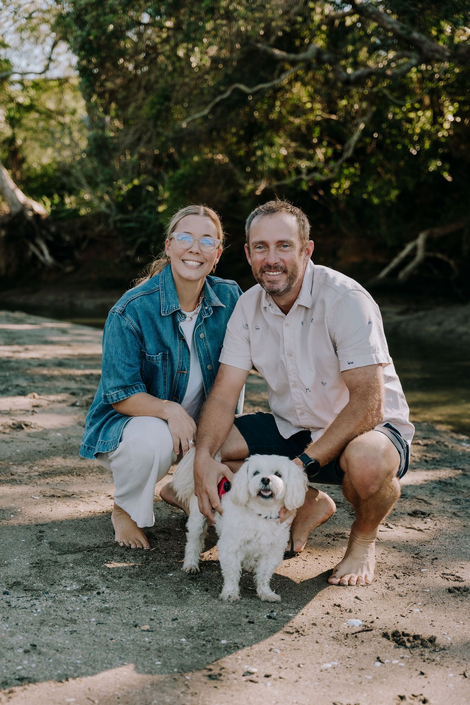A couple crouches on a sandy beach path with their small white fluffy dog in a natural outdoor setting.