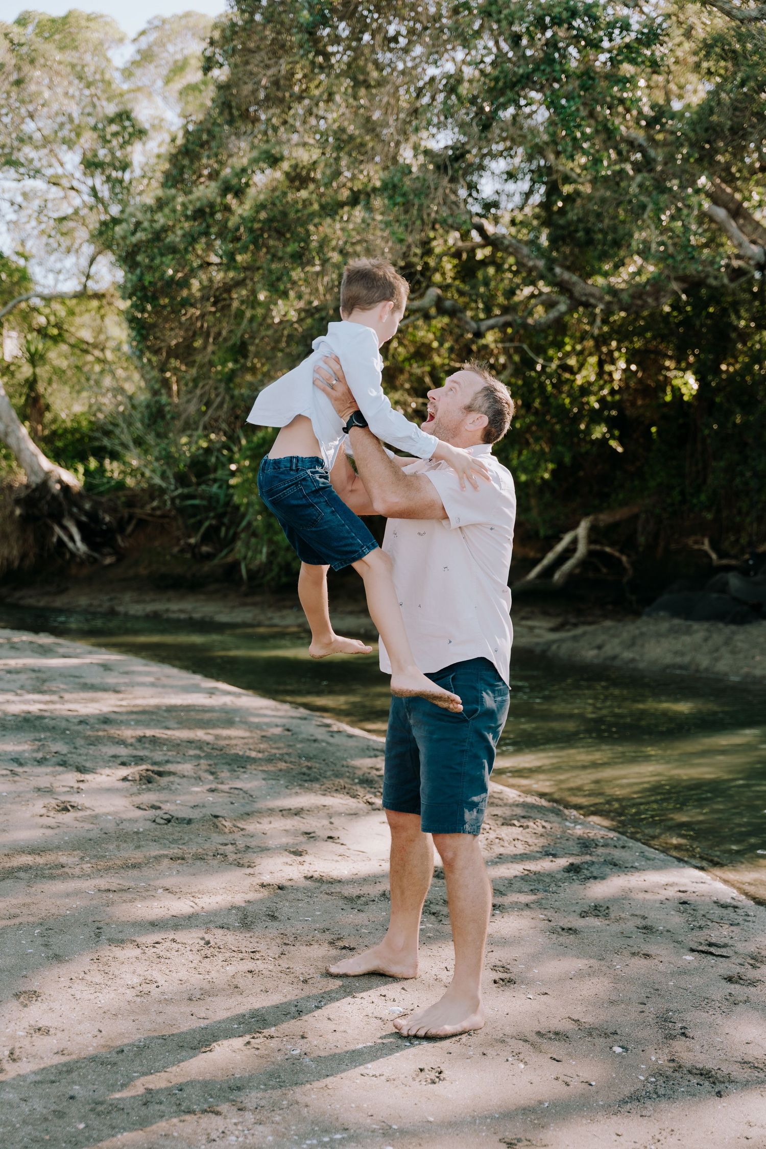 A tender moment captured at a sunny beach setting shows joyful play and lifting between two people on sandy ground.