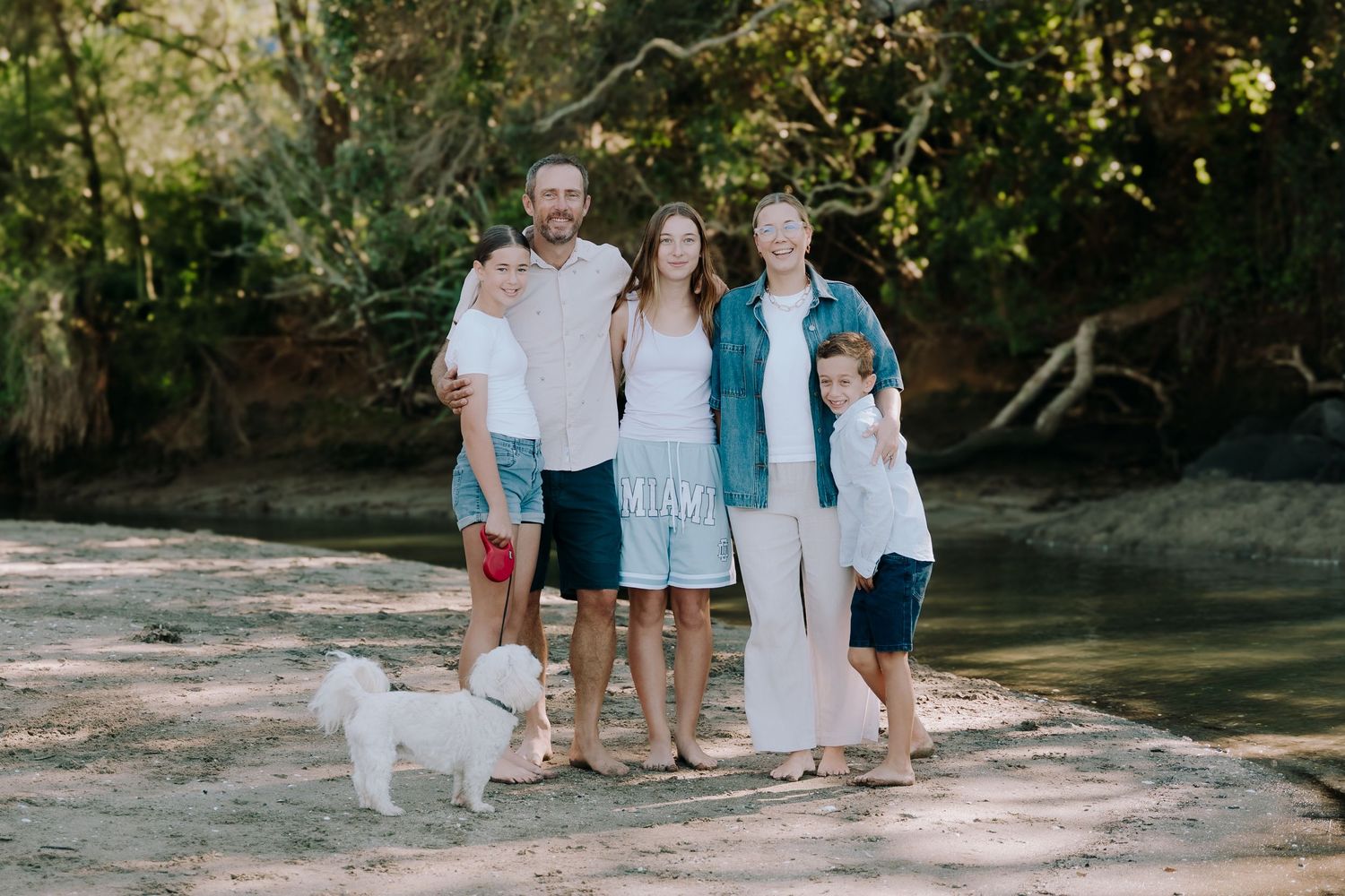 A family stands together outdoors on a sunny day with their small white dog, surrounded by lush greenery.