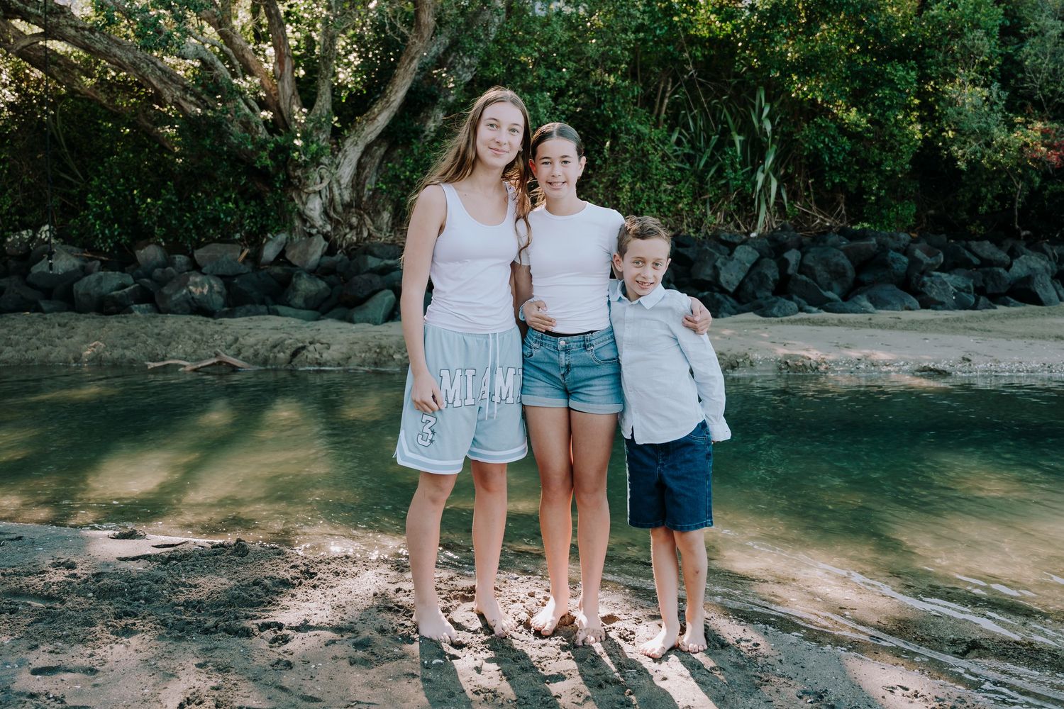 Three people standing together in front of a wooded area near a peaceful stream on a sunny day.