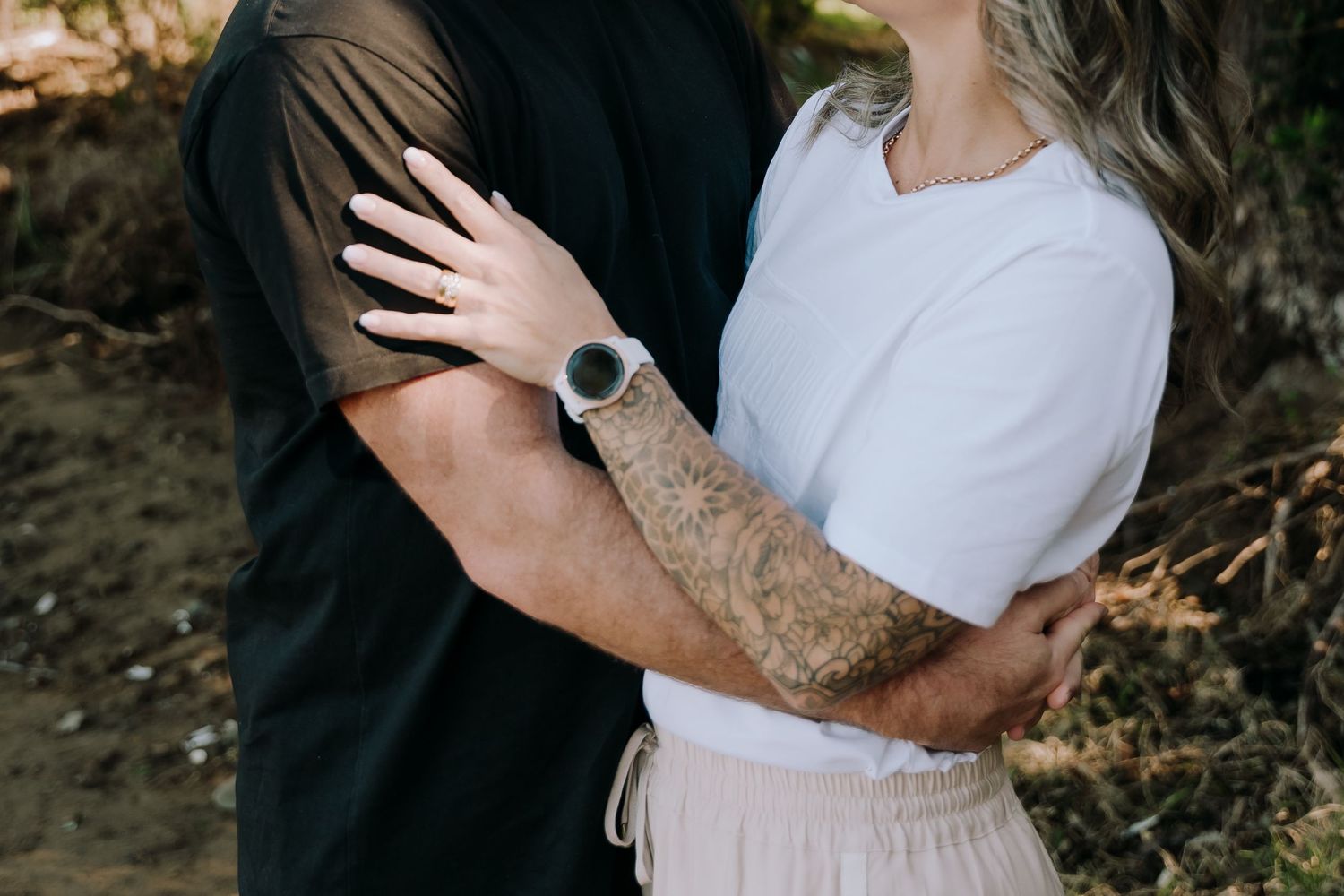 Couple embraces intimately with tattooed arms visible, wearing black and white tops against natural outdoor backdrop.