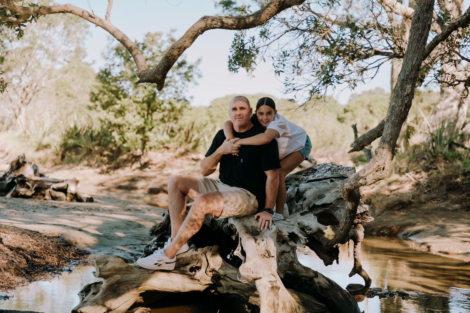 Two people sitting together on a fallen log by a peaceful creek in nature with sunlight filtering through trees.