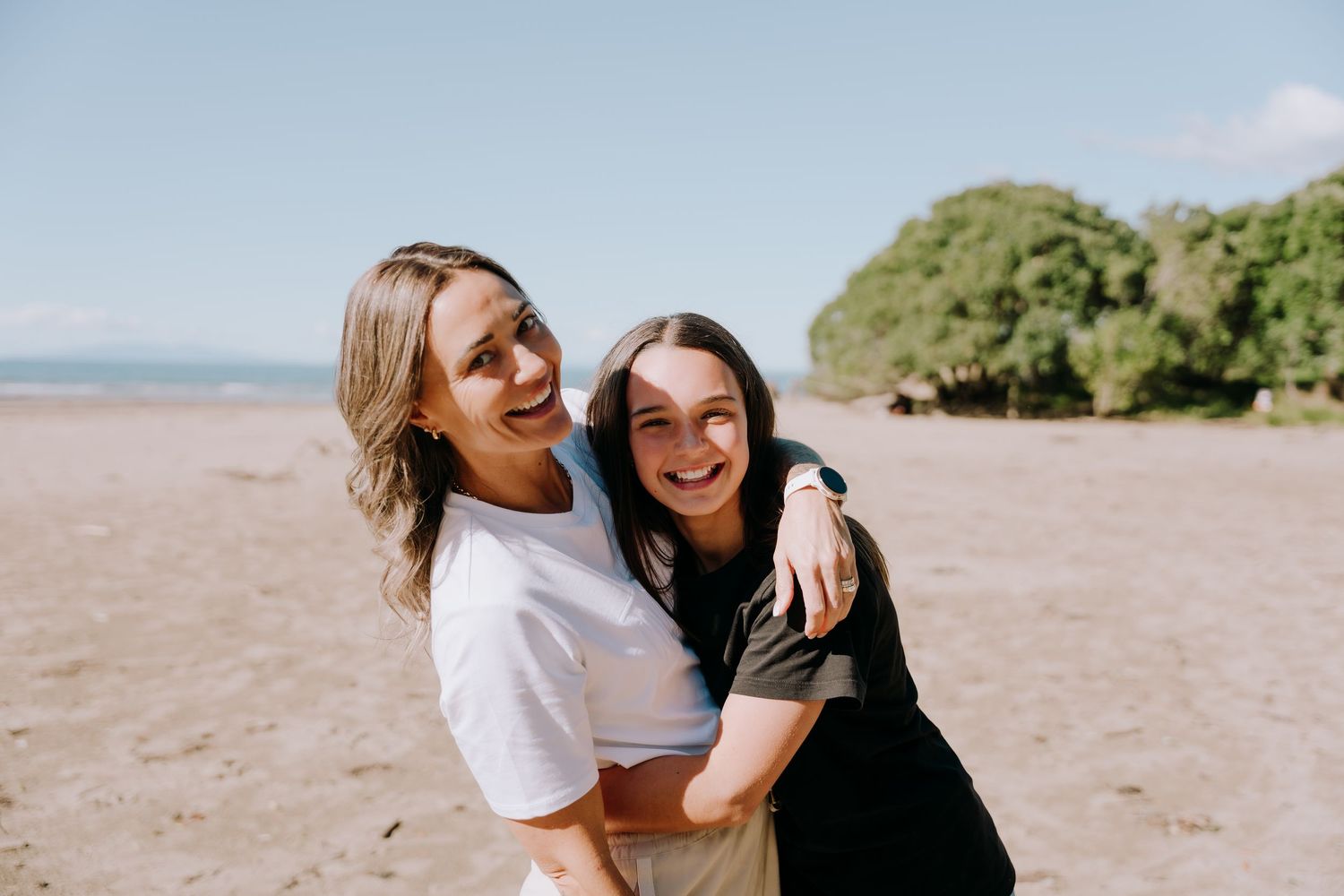 Two friends laughing and embracing on a sunny beach with trees in the background.