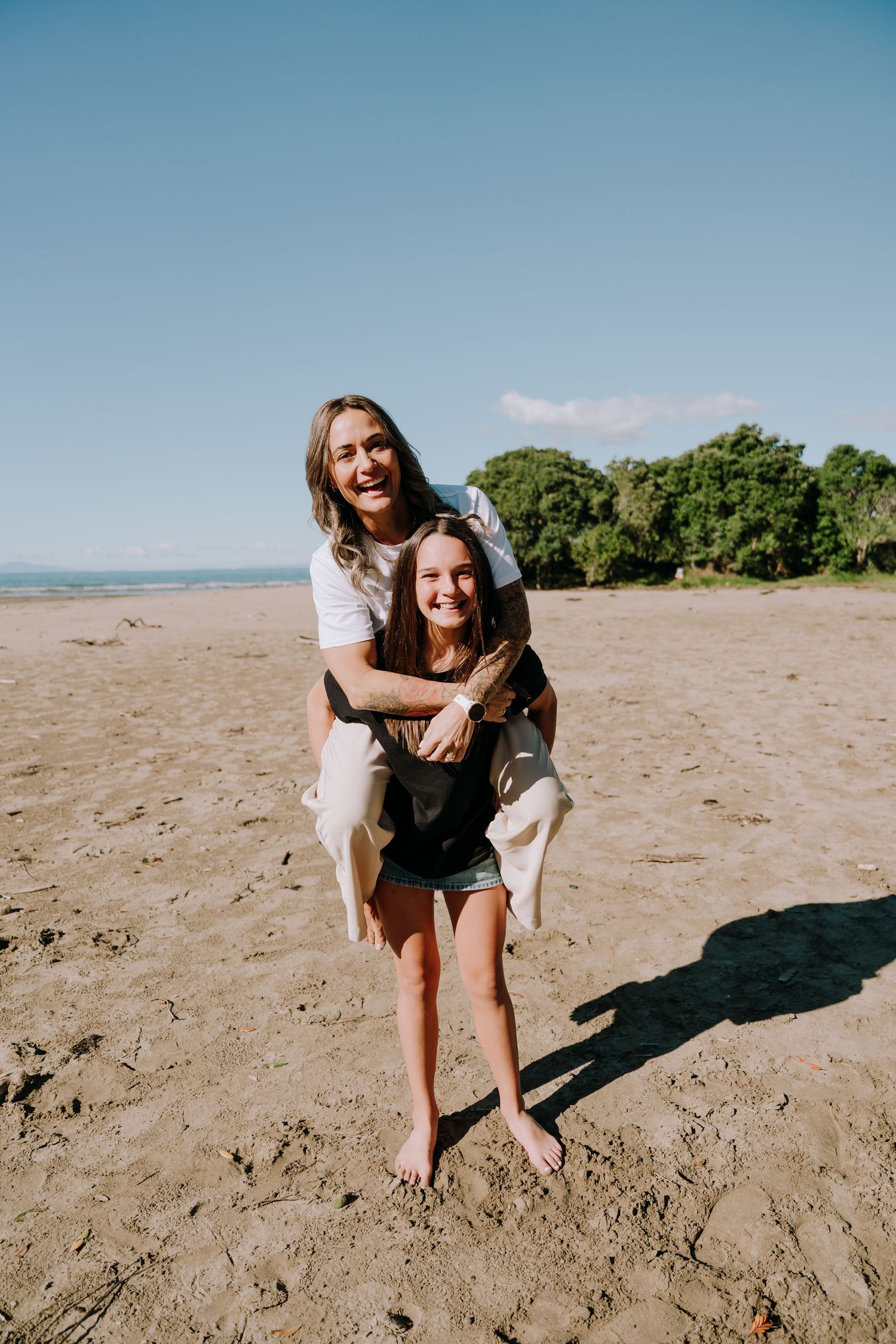 Two friends enjoying a playful piggyback ride on a sunny beach with trees and blue sky in the background.