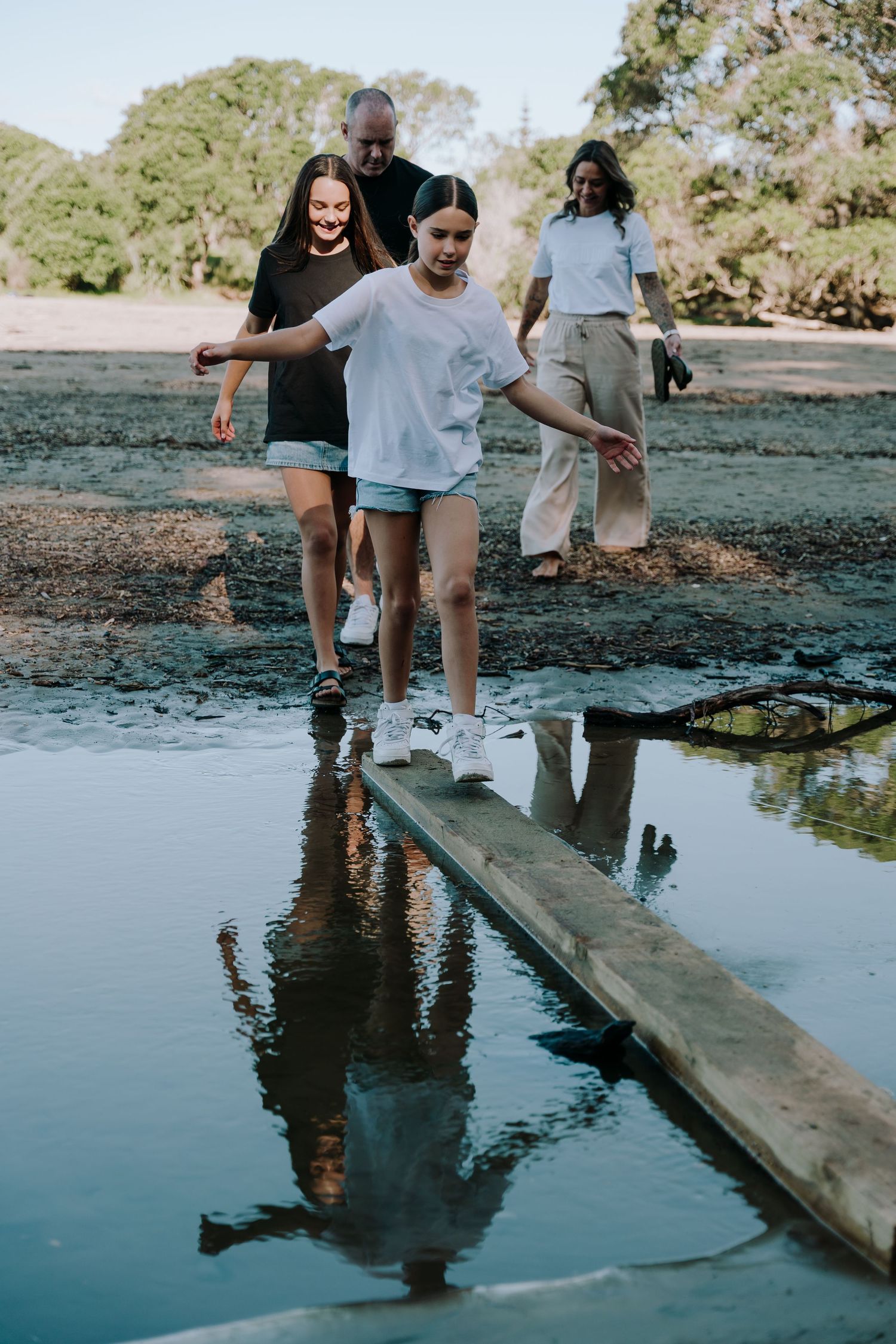 A group of friends balancing and walking on a wooden plank over a calm pond reflecting the scenic natural surroundings.
