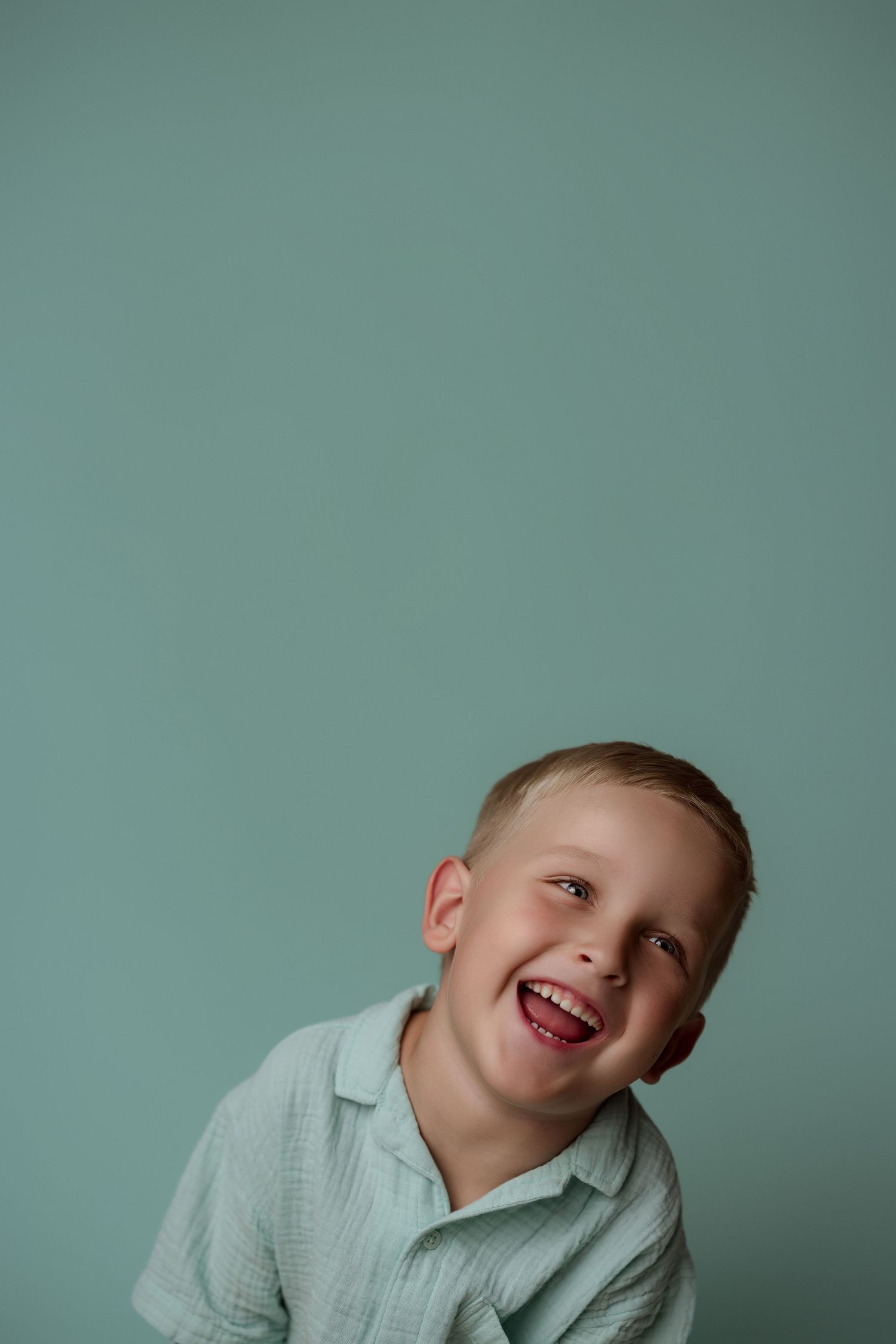 A child wearing a light colored shirt tilts their head playfully against a teal colored background.