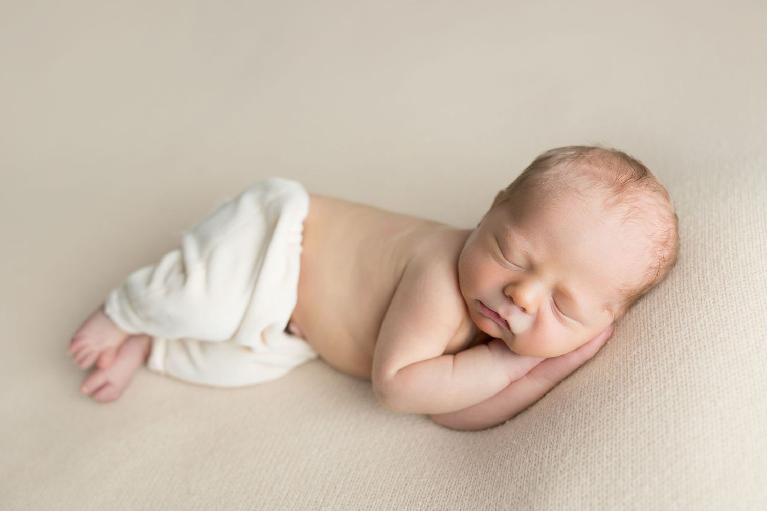Peaceful sleeping newborn baby wrapped partially in soft white fabric against beige backdrop.