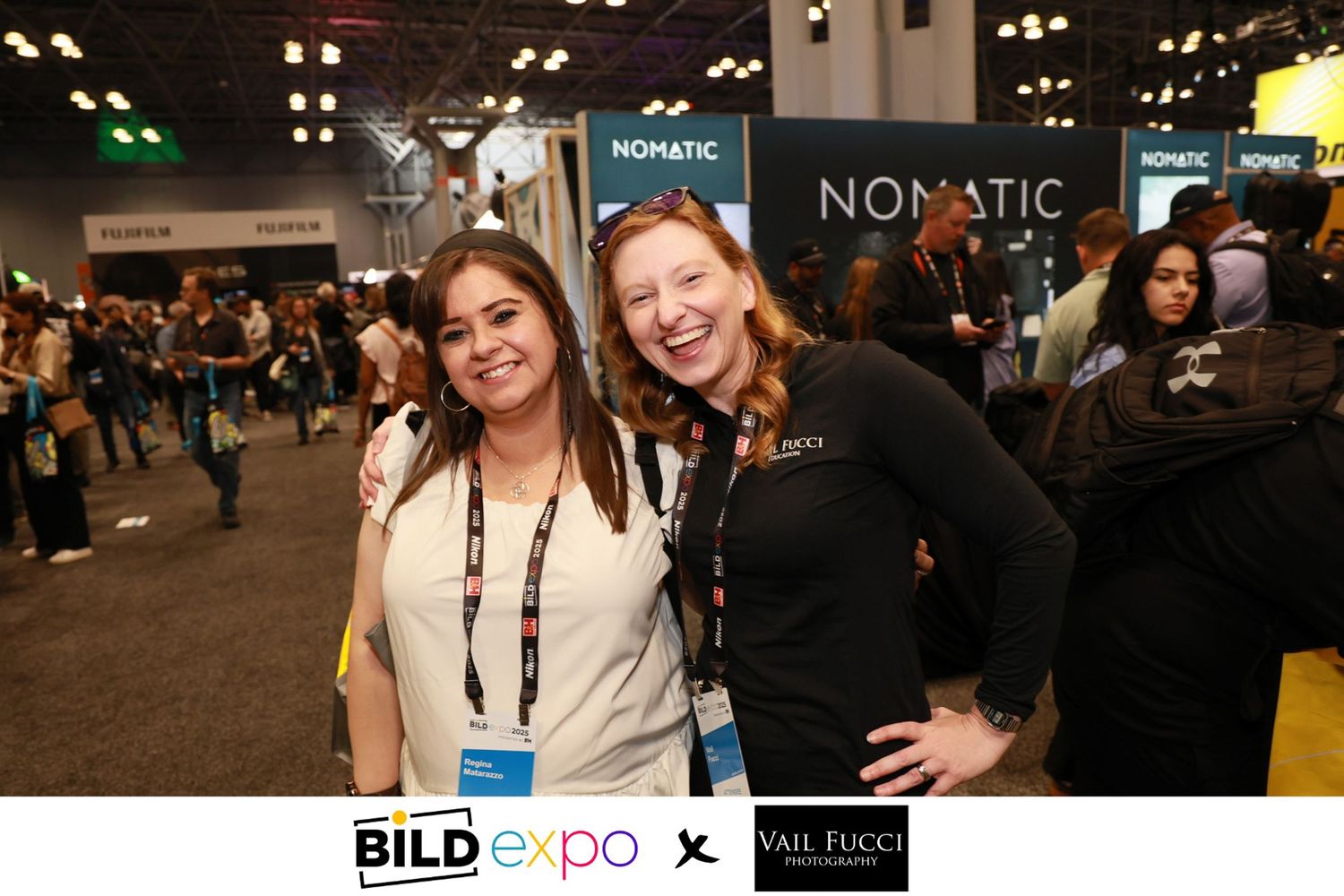Two attendees smile for a photo at a tech conference in front of the Nomatic exhibit booth.