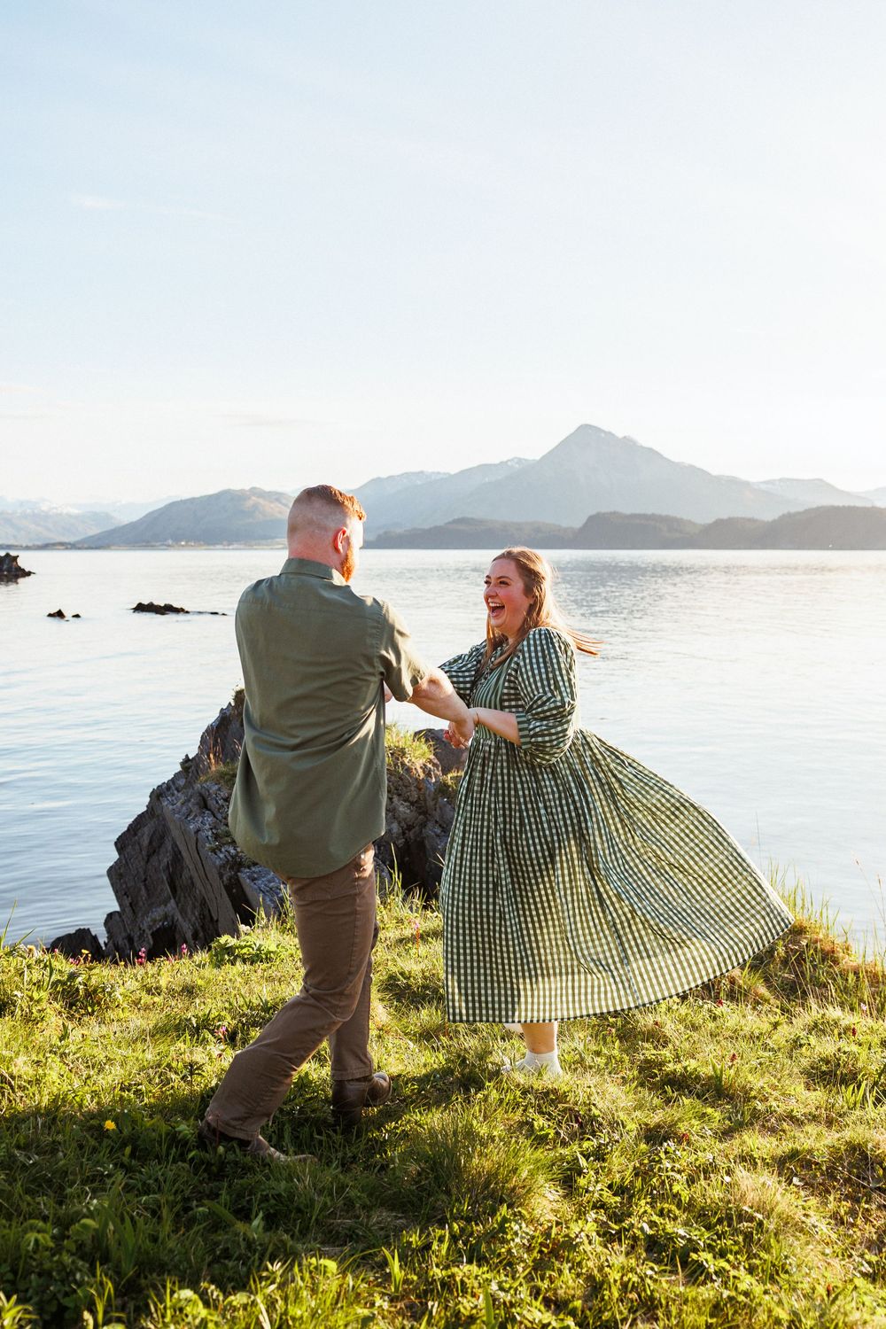 A couple shares playful moments on a grassy cliff overlooking a serene mountain lake at sunset.