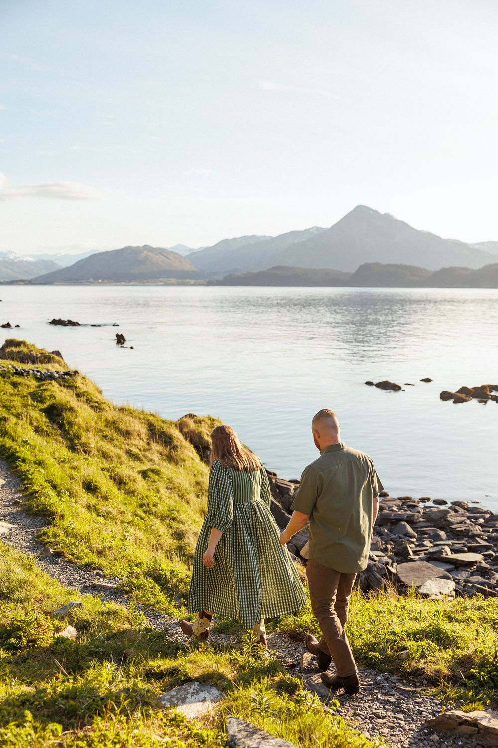 A pair walk along a coastal trail overlooking a serene mountain lake surrounded by rocky shores.