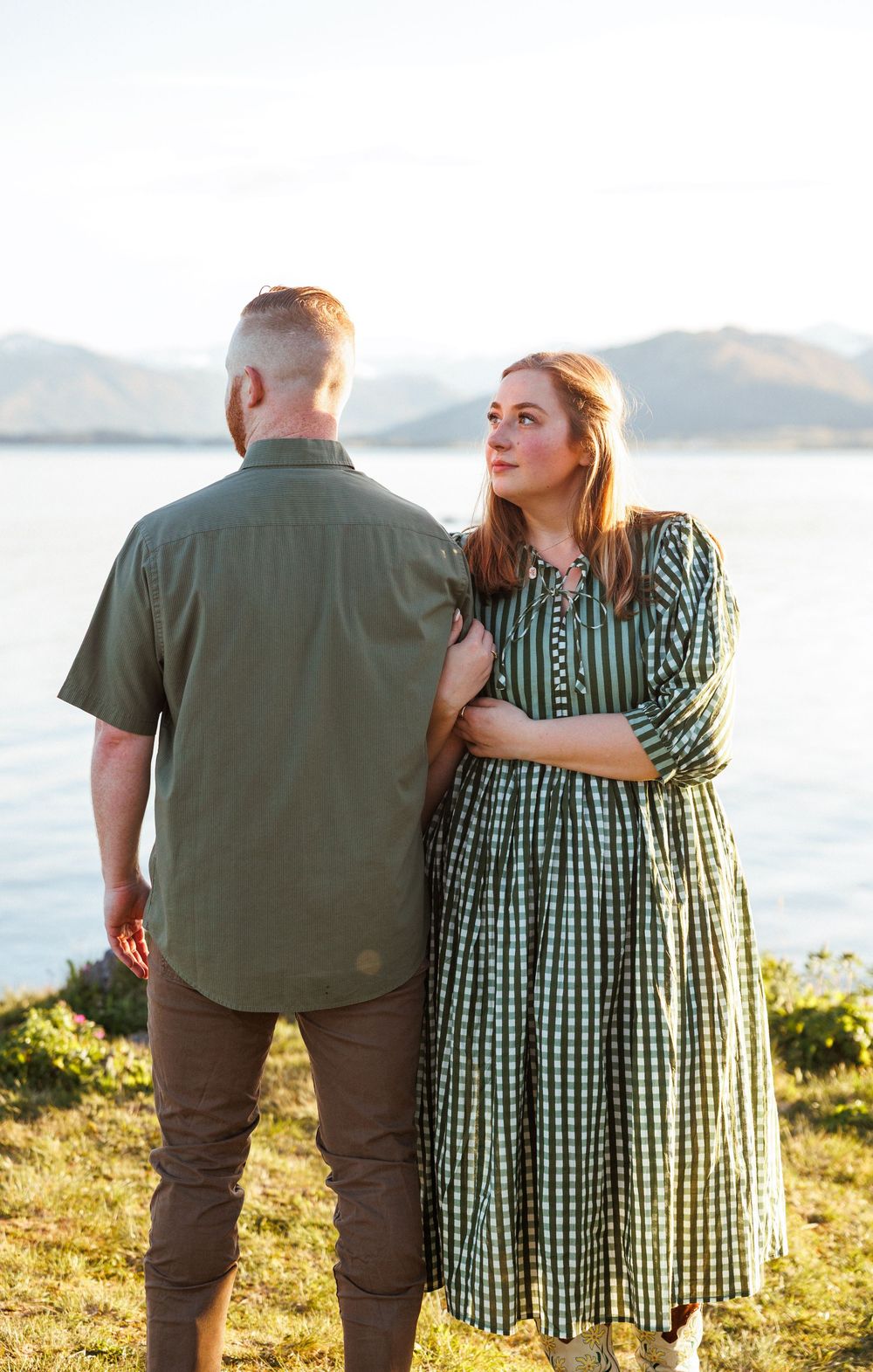A couple stands together beside a lake at sunset, with mountains in the background and warm evening light.