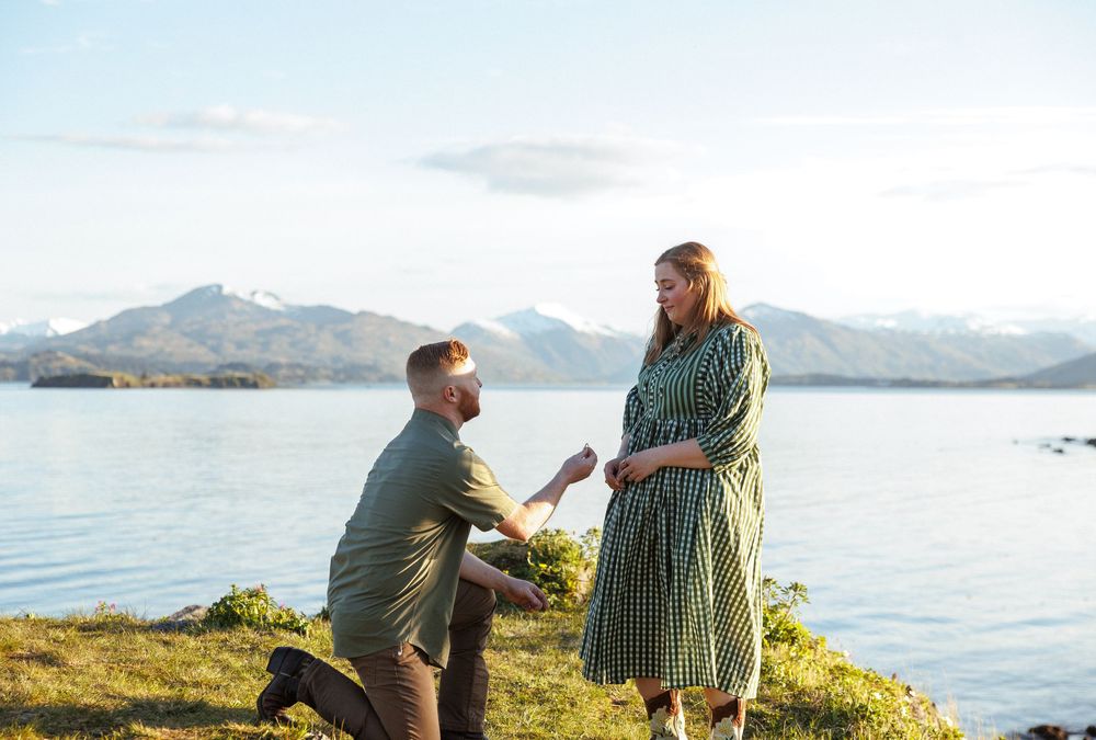 A romantic proposal moment takes place on a grassy hill overlooking a serene mountain lake on a sunny day.