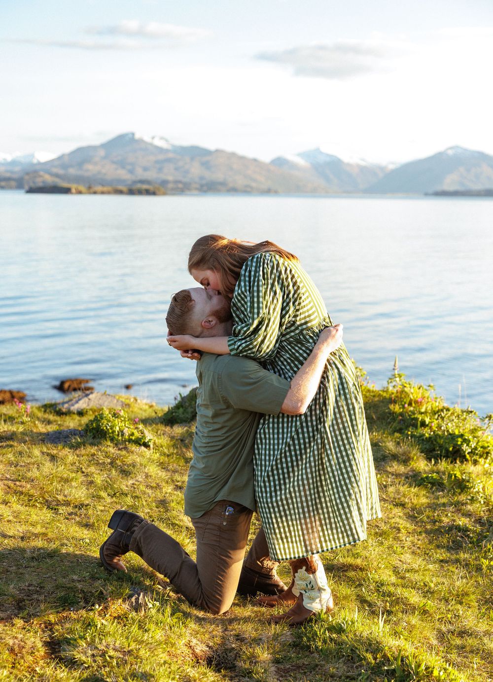 Two people embrace on a grassy hillside overlooking a serene lake and mountain landscape at sunset.