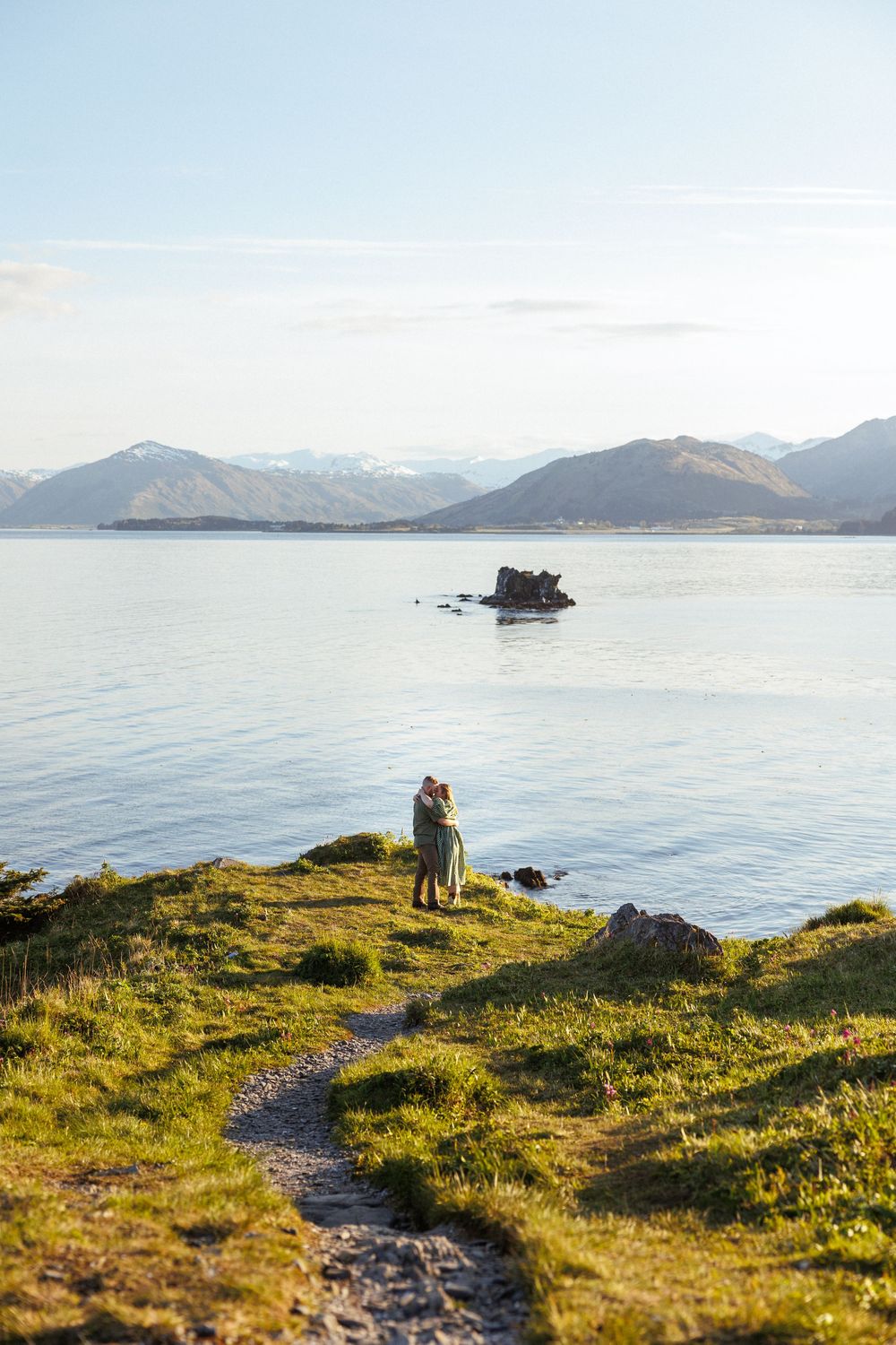 A couple stands together on a coastal hillside trail overlooking a tranquil mountain lake.