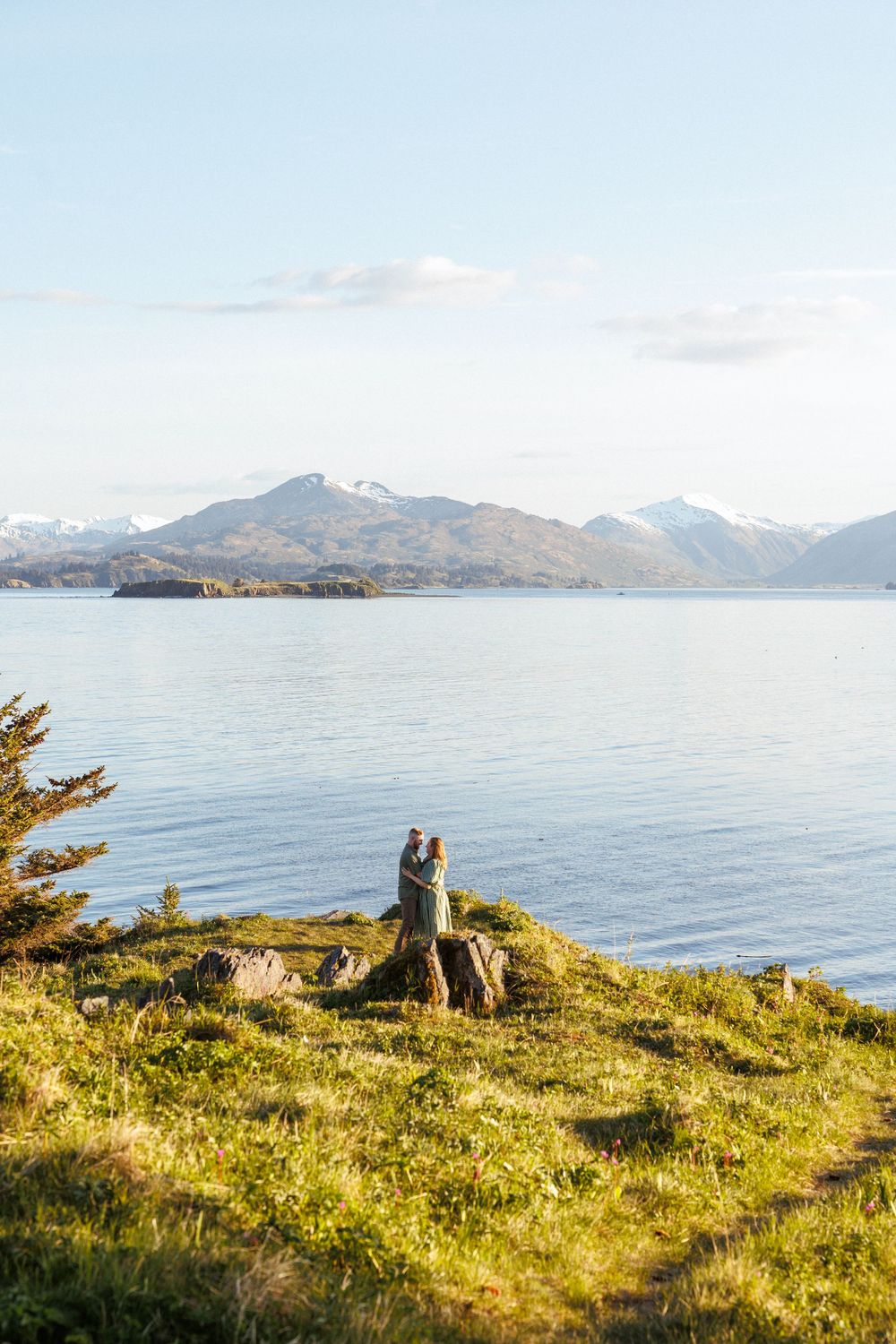 Two people sit together on a grassy cliff overlooking a scenic mountain lake with islands and snow-capped peaks in the distance.