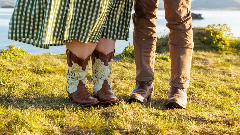 Close up of cowboy boots and casual shoes on grassy hillside with sun and lake in background.