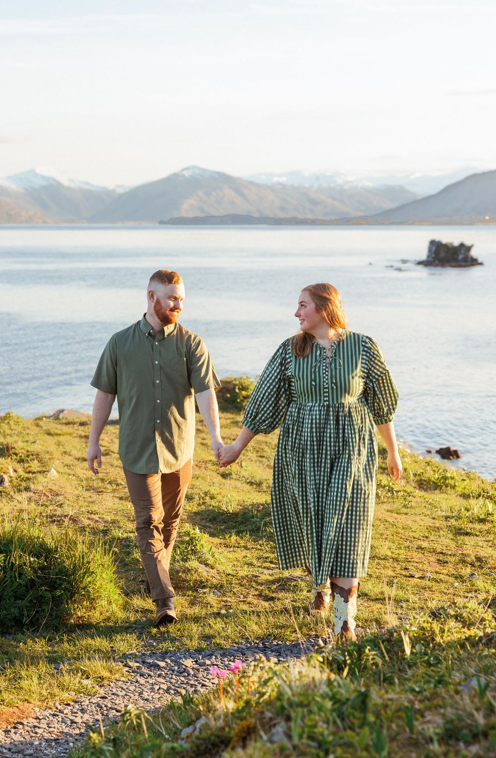 A couple strolls along a scenic coastal path with mountains and water in the background during golden hour.
