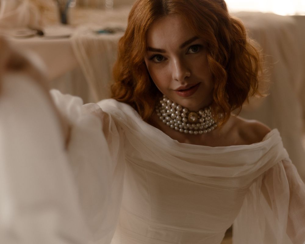 Model wearing pearl choker necklace and flowing white fabric in warm-toned portrait photography.