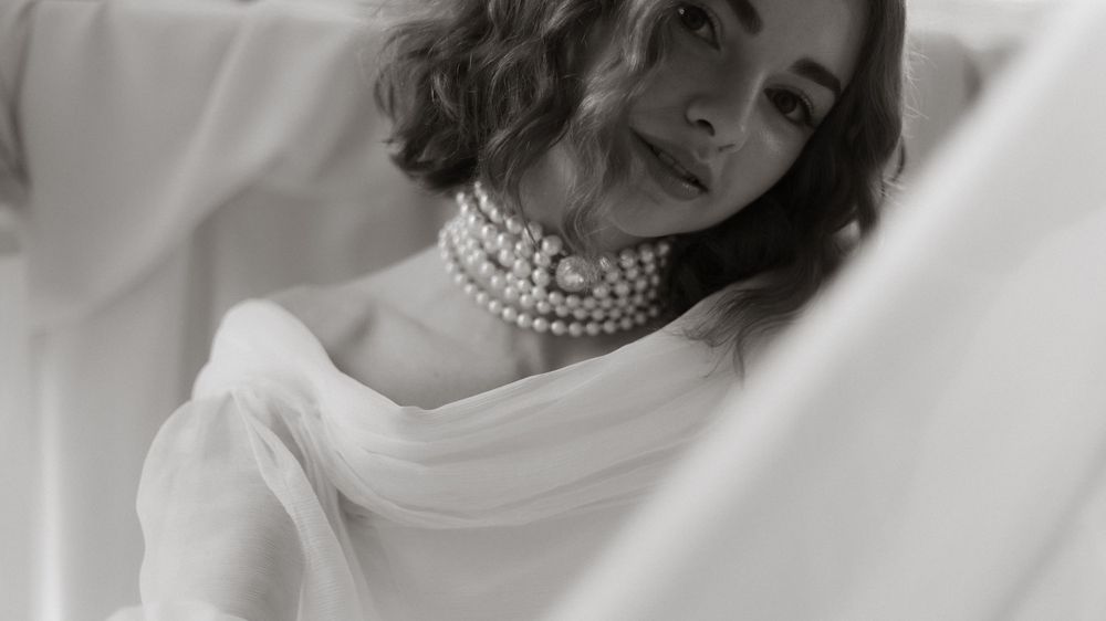 Black and white artistic portrait showing elegant multi-strand pearl necklace and white fabric draping against light background.