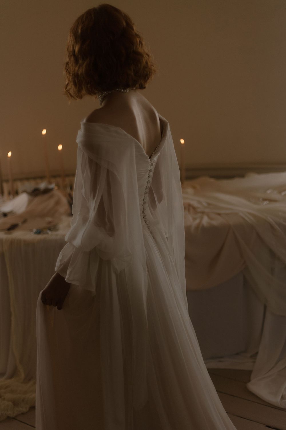 Back view of a person in a flowing white wedding dress with delicate buttons standing in a dimly lit room.