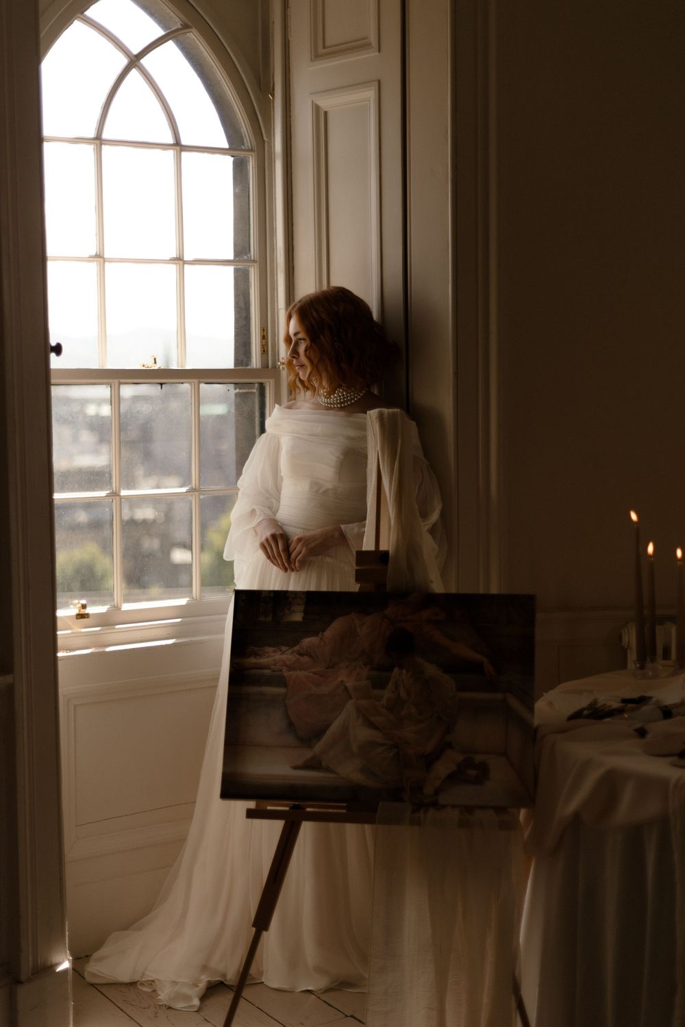 Figure in a white dress stands by an arched window with an art easel in a moody, candlelit interior setting.