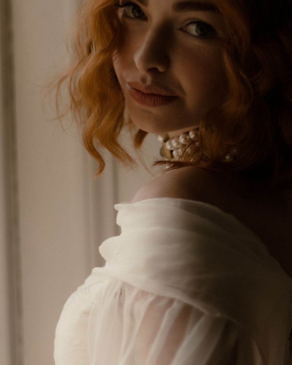 An artful portrait in soft lighting shows a figure in a delicate white blouse with flowing red hair against a light background.