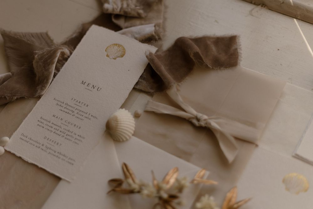 Wedding invitation with menu card displayed alongside delicate seashells and dried flowers in warm neutral tones.