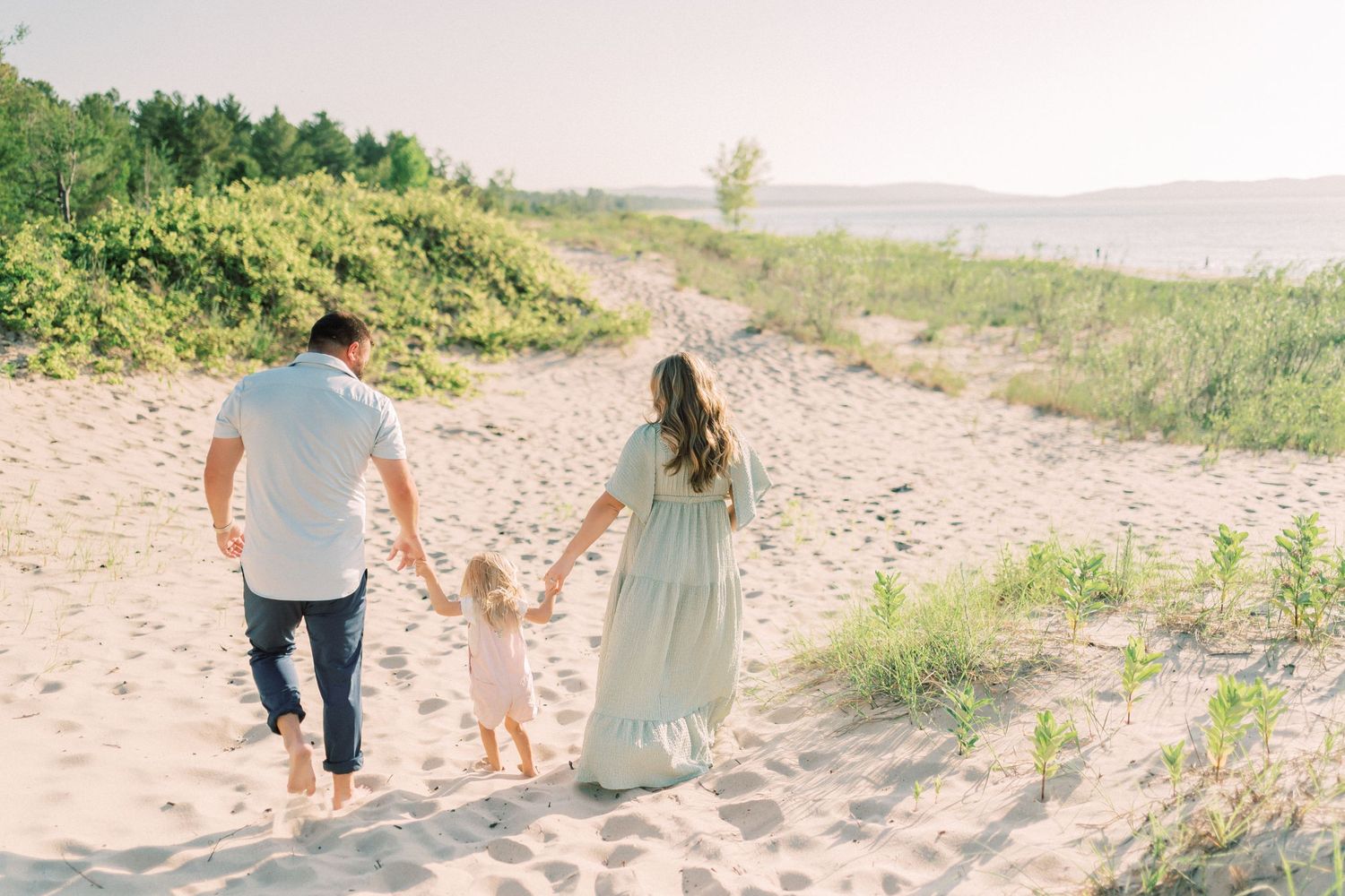 Three people walk together holding hands on a sandy beach path surrounded by dunes and coastal vegetation.