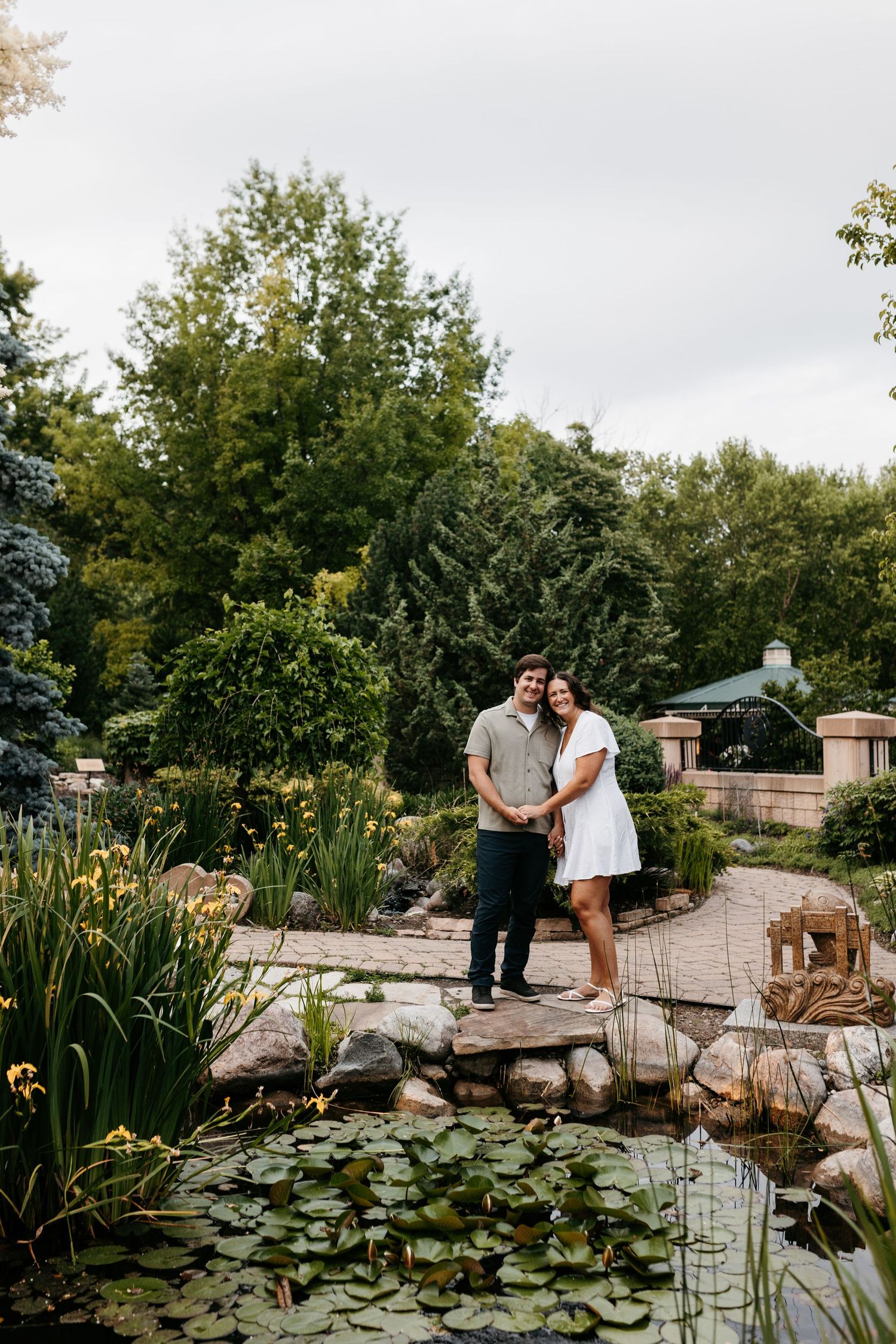 A couple stands on a stone bridge over a lily pond in a lush garden setting with trees and flowers.