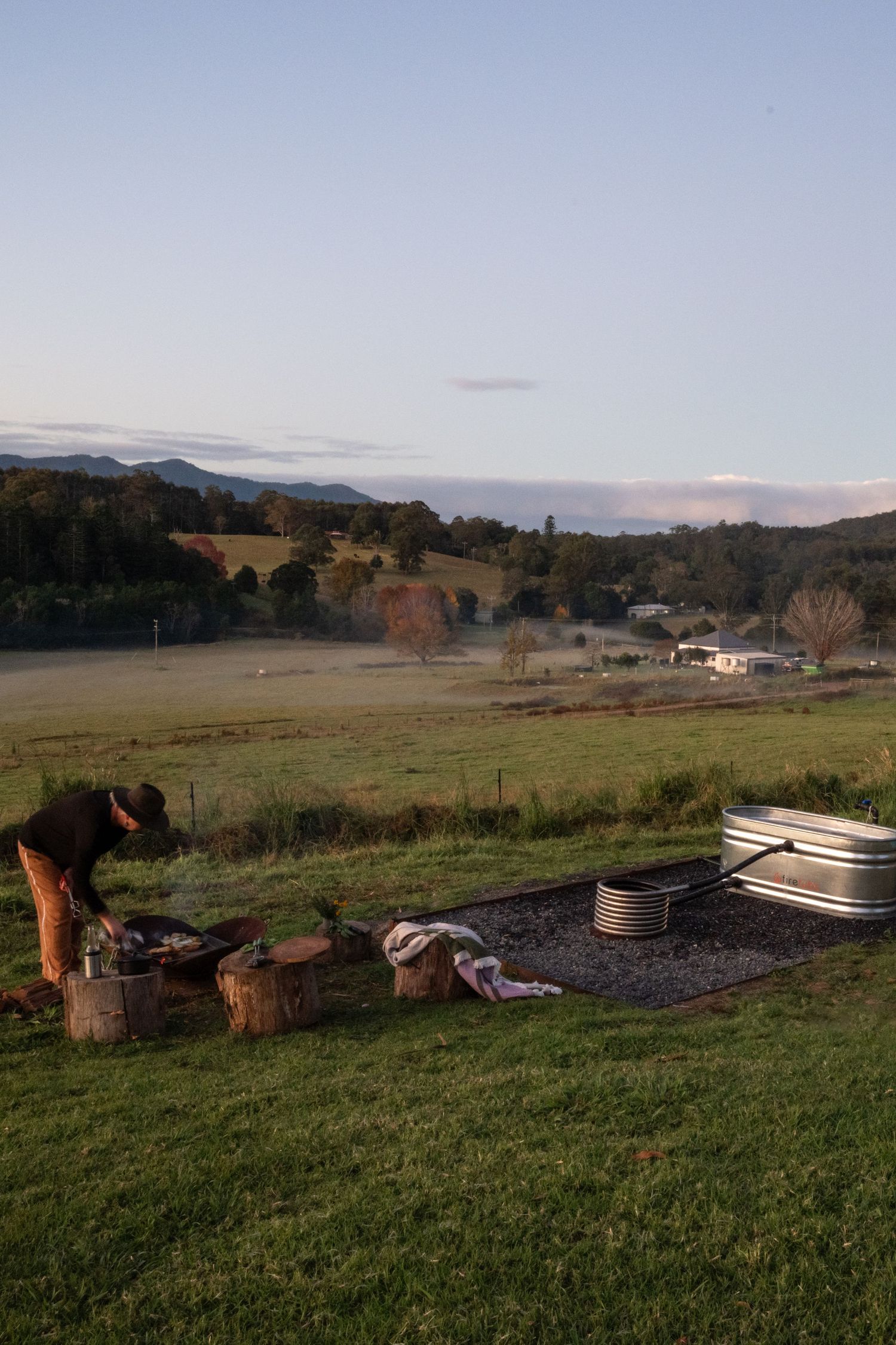 Misty rural landscape at dusk with camping setup including fire pit and cooking equipment on grassy hillside overlooking valley.