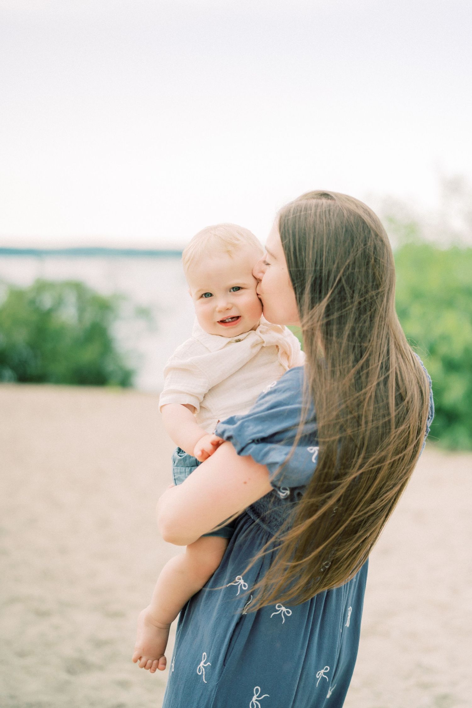 Parent in denim shirt holding and embracing small baby against beach backdrop in warm sunlight.