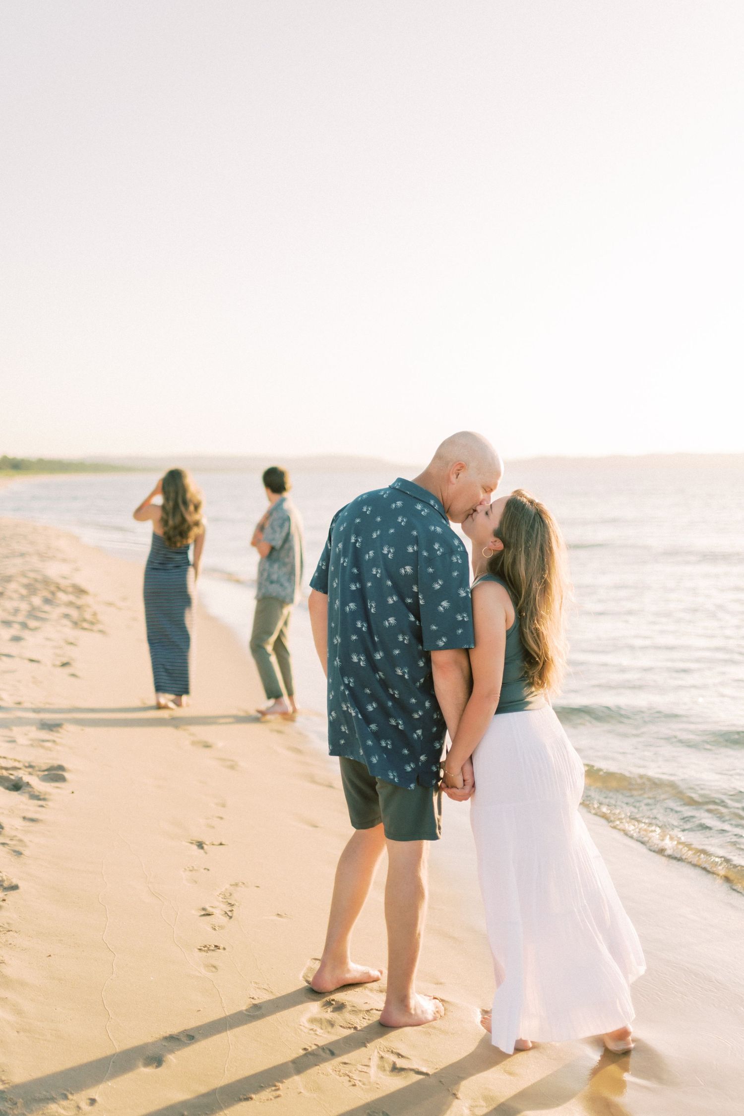 A couple strolls intimately along a sunny beach shoreline wearing white and navy blue outfits.