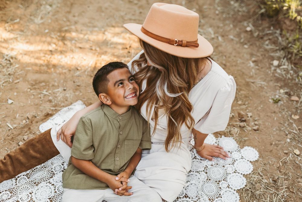 A woman in a white dress and brown hat sits on a lace blanket outdoors with a child wearing an olive colored shirt.