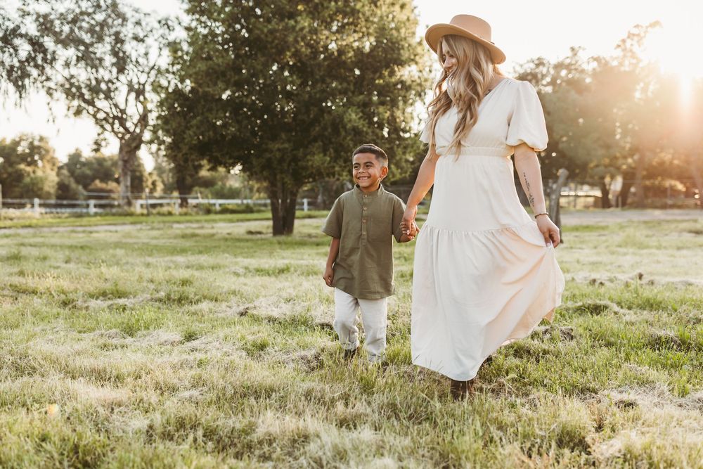 A parent and child walk hand in hand through a sunlit meadow at golden hour, wearing casual summer attire.