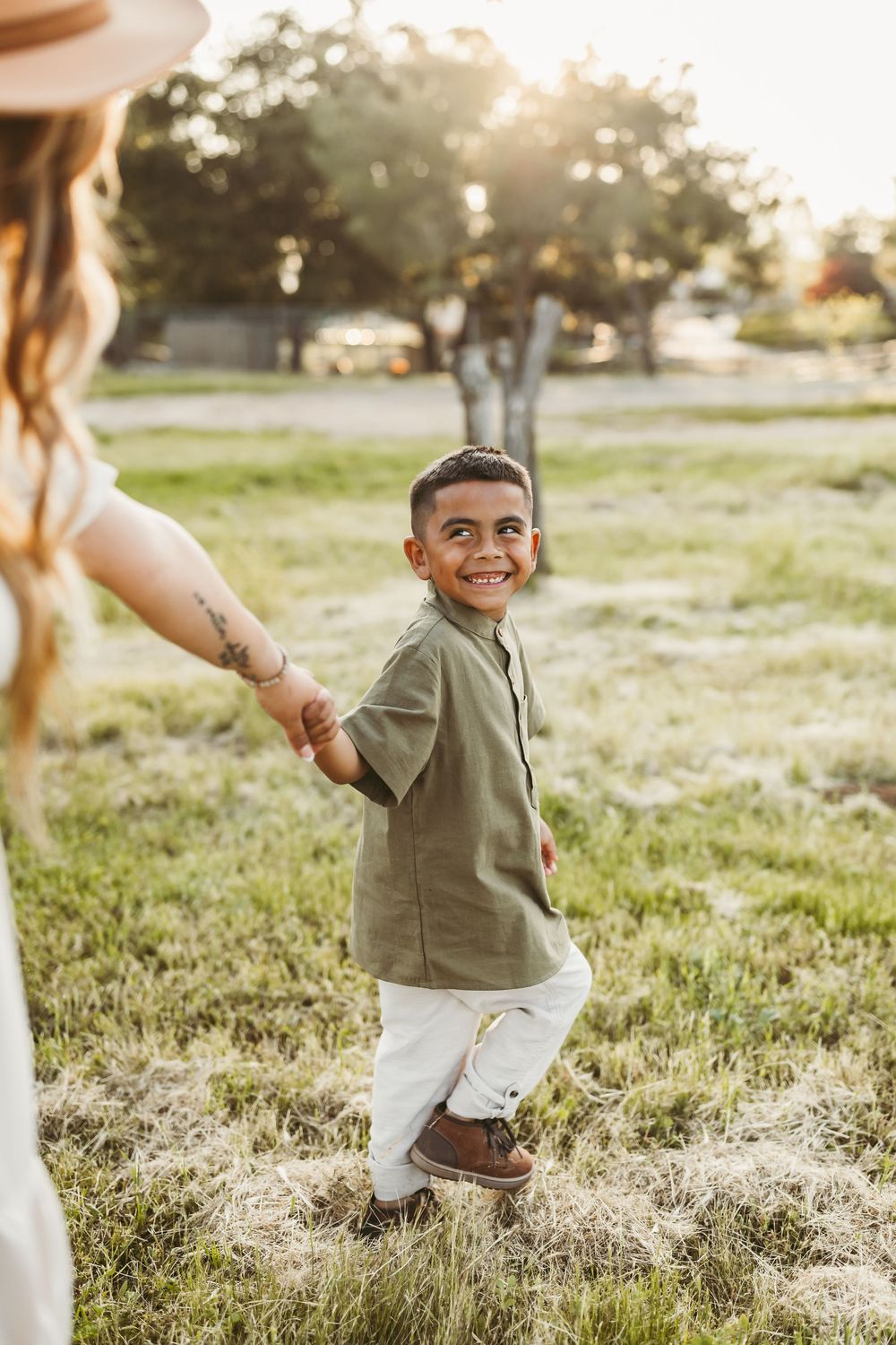 Young child in olive green shirt and white shorts holds hand with someone while playing in a sunny field at golden hour.