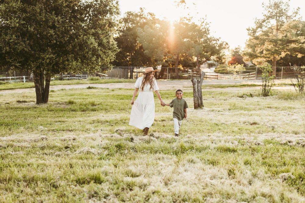Mother and son walks through sun-drenched field at golden hour creating dreamy silhouettes against the setting sun.