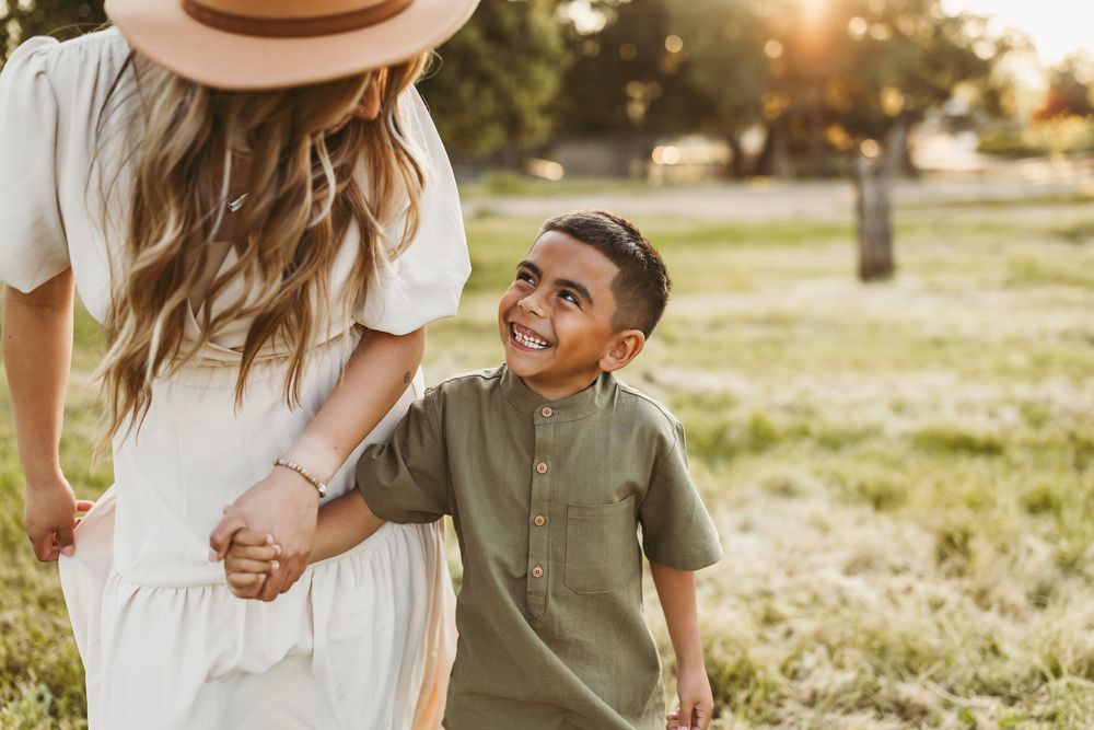 A mother and son hold hands while sharing a joyful moment together in a sunny park at golden hour.