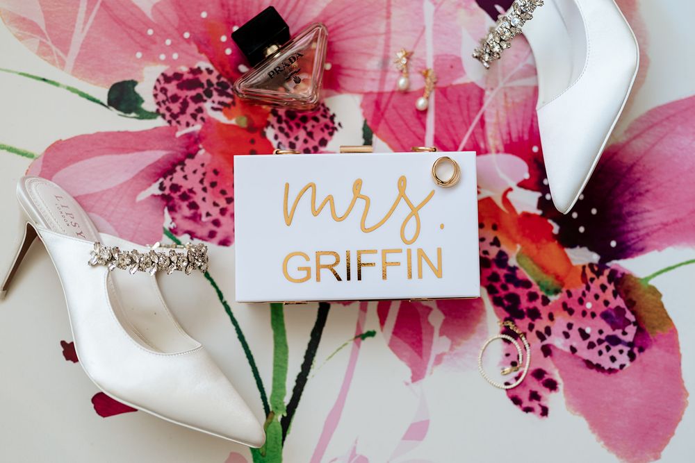 Wedding details with white heels, a 'Mrs Griffin' name card, and pink orchid flowers against a light background.