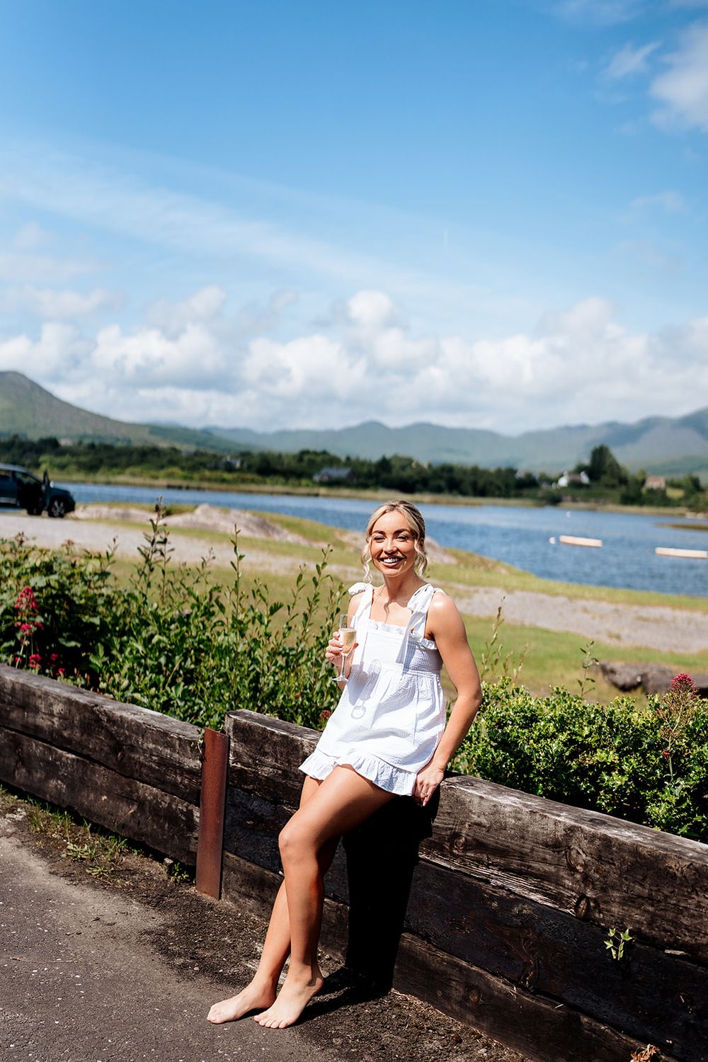 A person in white summer attire sits on a wooden barrier near a coastal bay with mountains in the background.