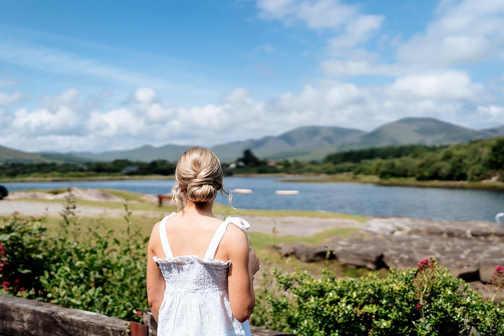Someone in a white dress overlooks a serene coastal bay with mountains and blue skies in Ireland.