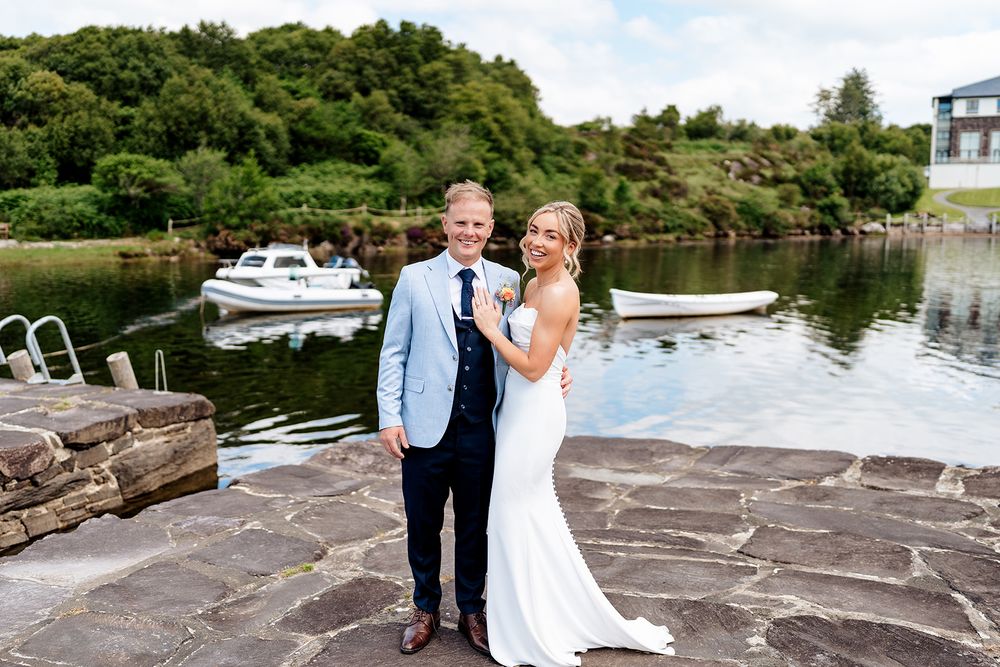 A couple in wedding attire stands on a stone dock by a lake with boats and green hills in the background.