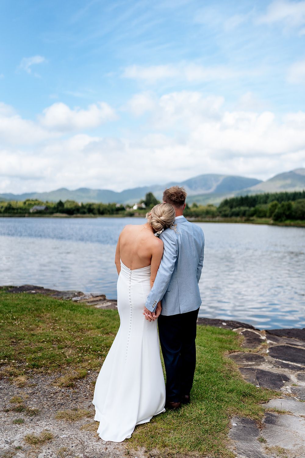 A couple in wedding attire embraces while looking out over a scenic mountain lake on their special day.