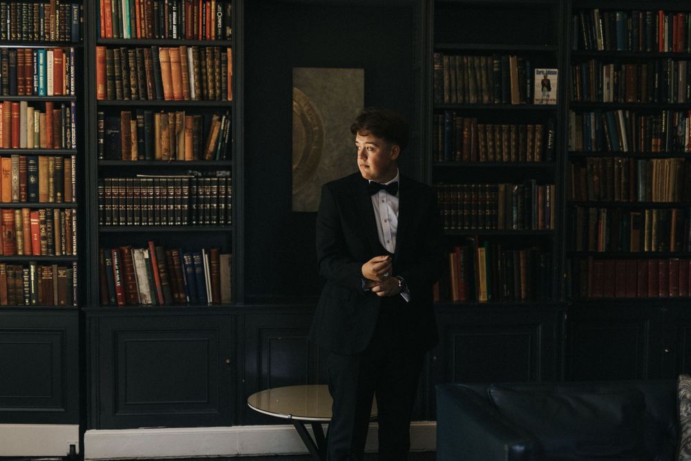 Person in black tuxedo standing against dark library wall filled with vintage books at Saltmarshe Hall - taken by Grimsby photographer.