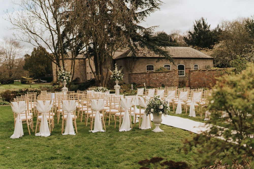 White fabric-draped chairs line a scenic outdoor wedding ceremony setup with trees and greenery at Saltmarshe Hall.