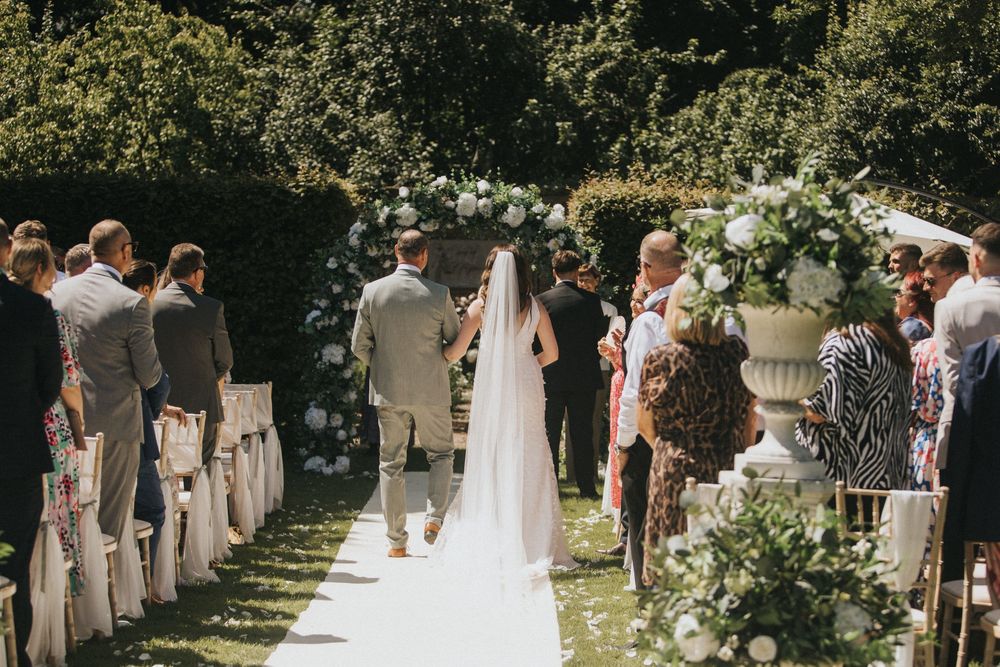 Wedding guests gather beneath trees in an outdoor garden ceremony during a sunny afternoon at Saltmarshe Hall - by Grimsby photographers.